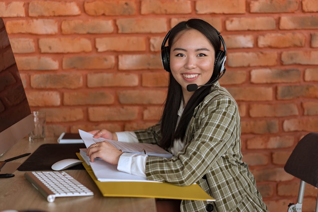 Asian woman working as a call center agent, smiling at desk with documents and headset.