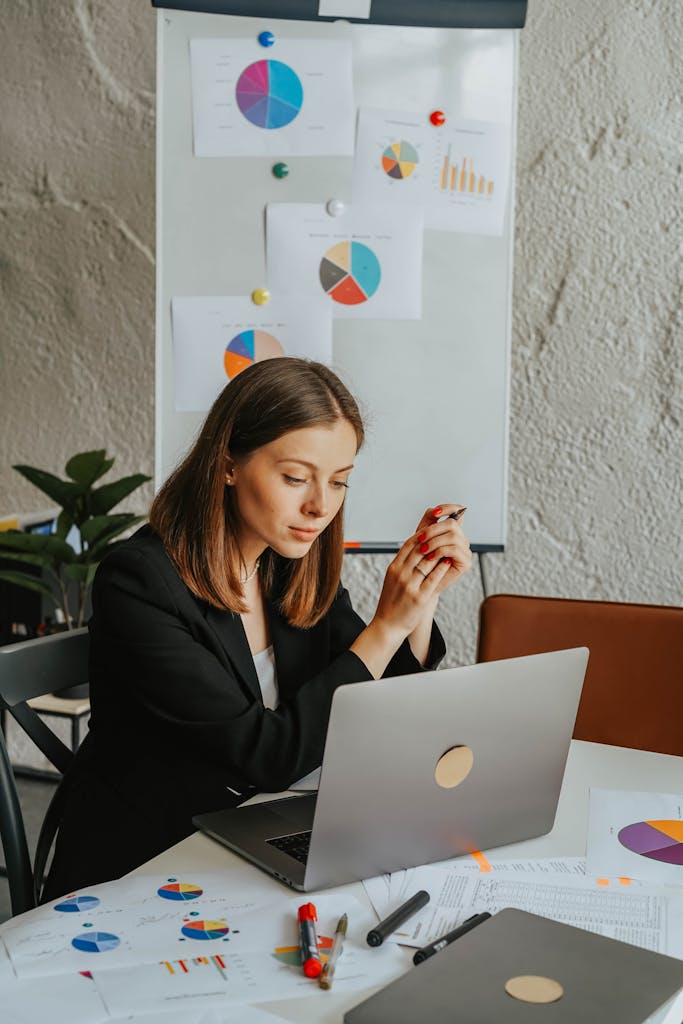 Businesswoman reviewing charts at a laptop in an office setting with presentations behind her.