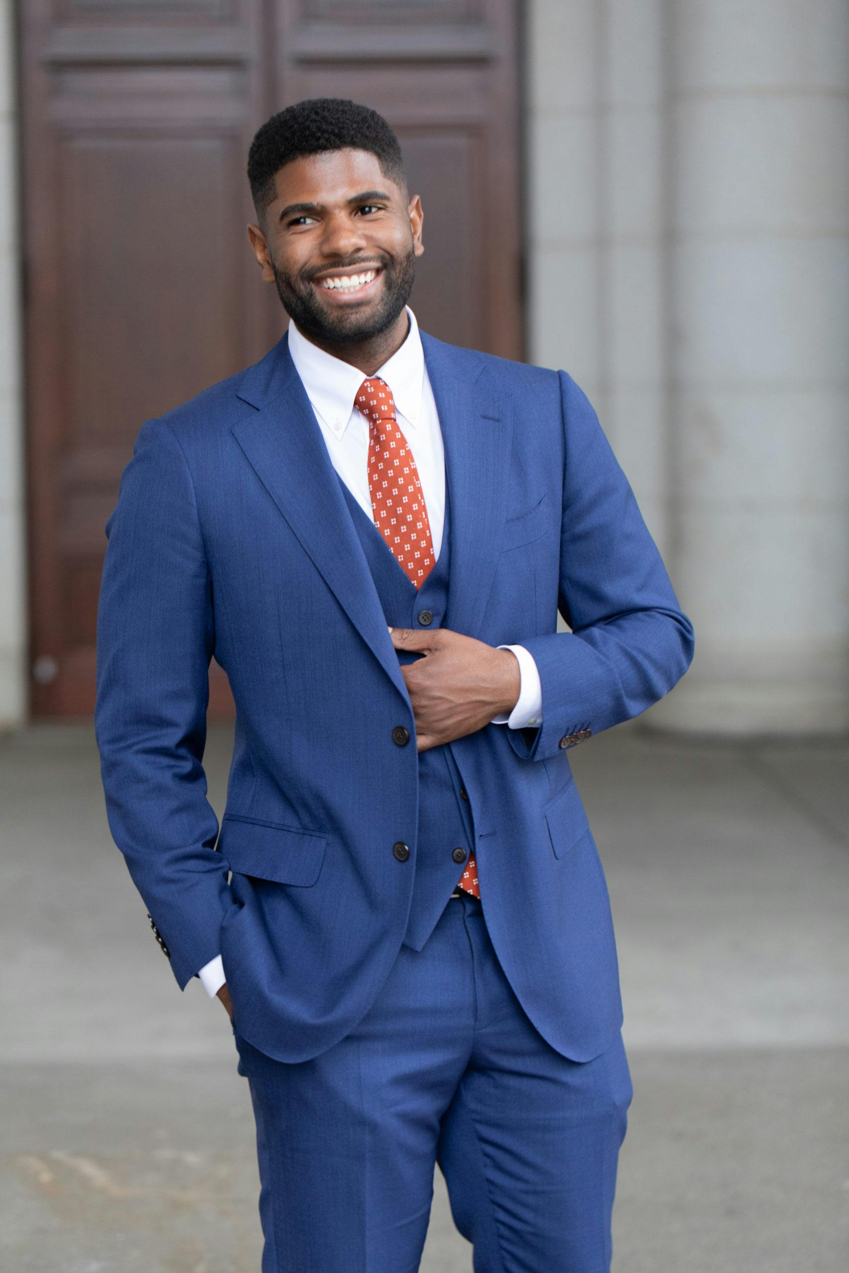 Smiling businessman in a blue suit posing comfortably outdoors with an elegant demeanor.