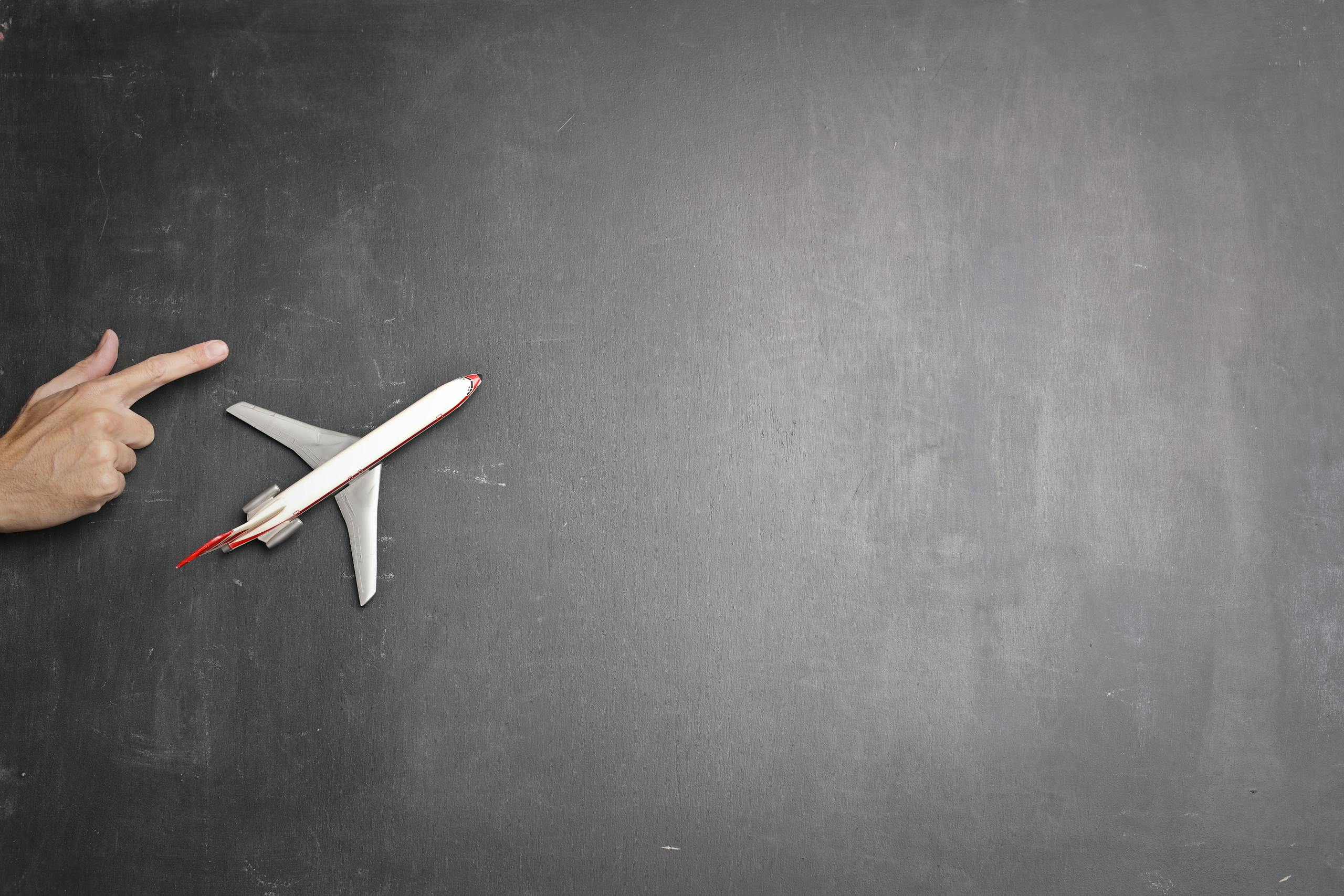 Conceptual image of a hand pointing towards a toy plane on a chalkboard background, symbolizing travel and direction.